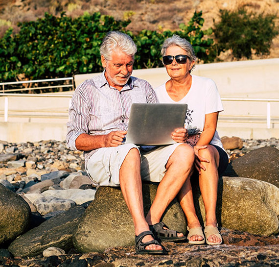 senior couple sitting on beach rocks on their laptop researchign medicare parts explained houston tx